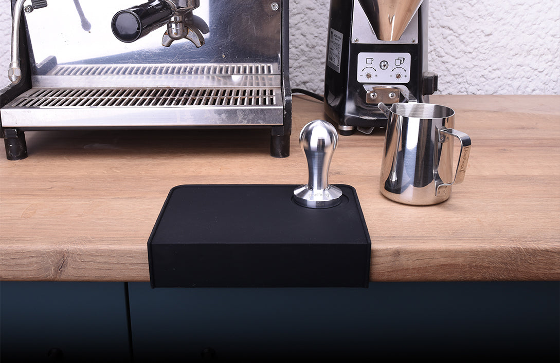 black silicon tamping mat with tamper and milkpitcher next to coffee machine and grinder. barista & espresso tools by joefrex